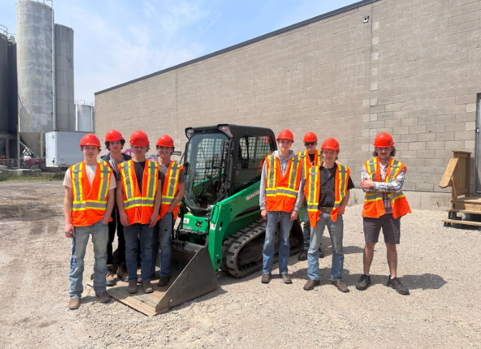 A group of students wearing high vis vests and safety helmets standing outside next to a digger.