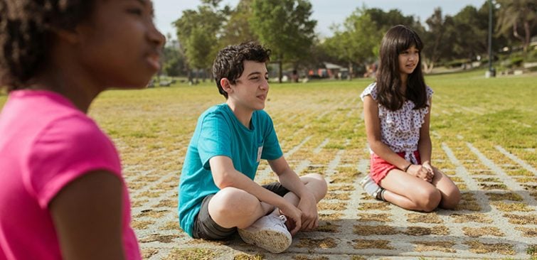 Three kids sit on the grass outside on a sunny day